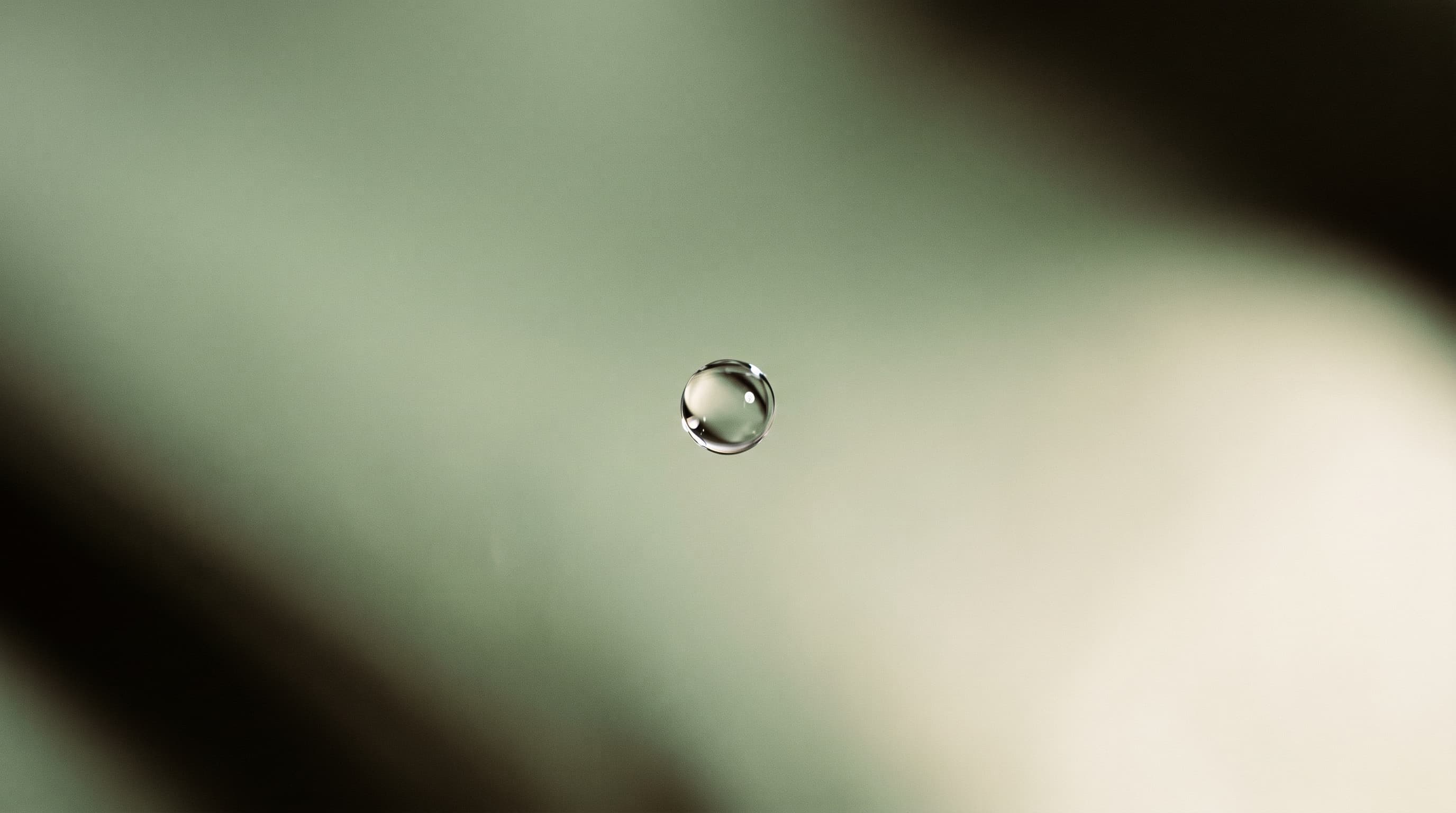 A single water droplet caught against a soft sage background